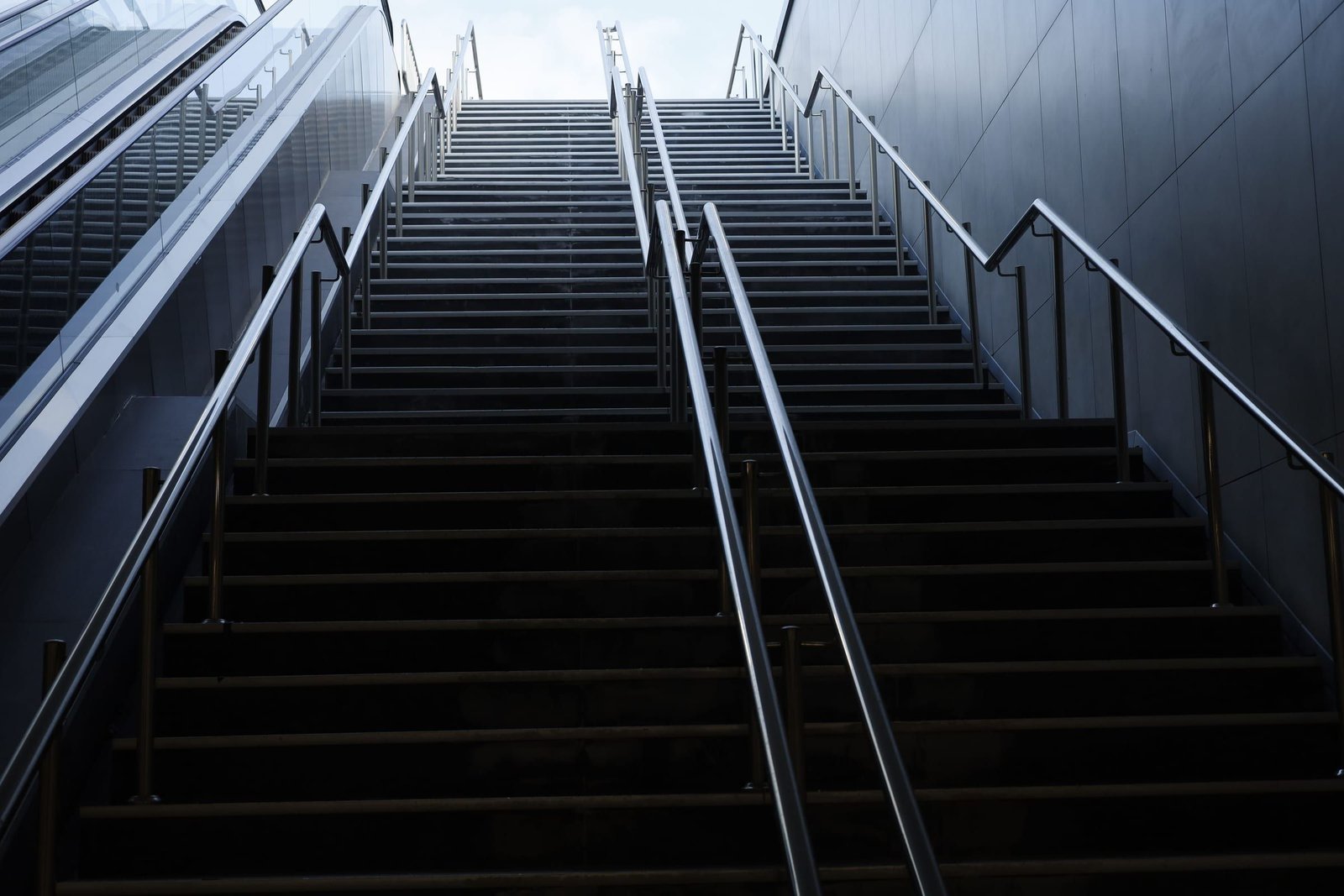 Long escalier urbain qui monte vers la lumière, encadré de rampes métalliques.